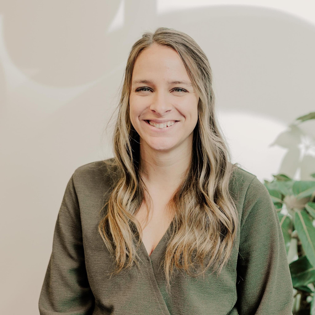 Headshot of Amber Darsch. Amber is a white woman in her mid-thirties with wavy blonde/brown hair. She is seated smiling with a plant in the background.