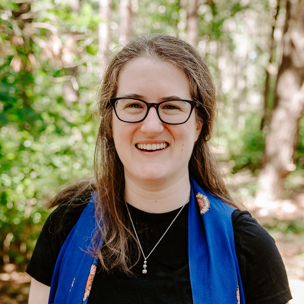 Headshot of Ellen Mason. Ellen is a white woman in her mid-thirties with wavy brown hair and glasses. She is outside in the woods, smiling.